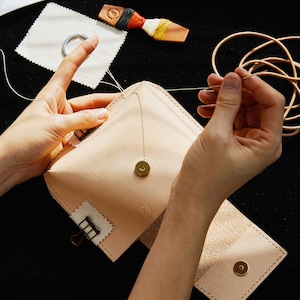 May include: A person is hand-stitching a light brown leather pouch with a gold button and a metal clasp. The pouch is open and the inside is lined with a light brown fabric. There is a spool of thread and a piece of leather on the table.