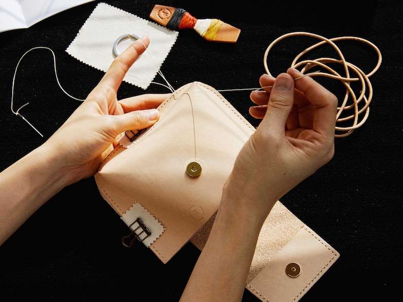 May include: A person is hand-stitching a light brown leather pouch with a gold button and a metal clasp. The pouch is open and the inside is lined with a light brown fabric. There is a spool of thread and a piece of leather on the table.