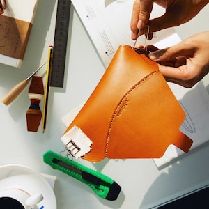 May include: A person is sewing a brown leather pouch with a white fabric lining. The pouch has a curved shape and a small label that says "Atelier." The person is using a needle and thread to sew the pouch together.