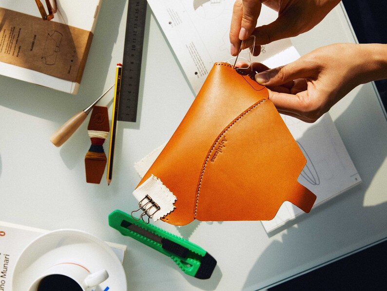 May include: A person is sewing a brown leather pouch with a white fabric lining. The pouch has a curved shape and a small label that says "Atelier." The person is using a needle and thread to sew the pouch together.