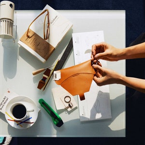 May include: A person working on a leather craft project. They are holding a brown leather piece and a pattern on a white surface. There are other tools and supplies on the table, including a book titled "Williams" and a cup of coffee.