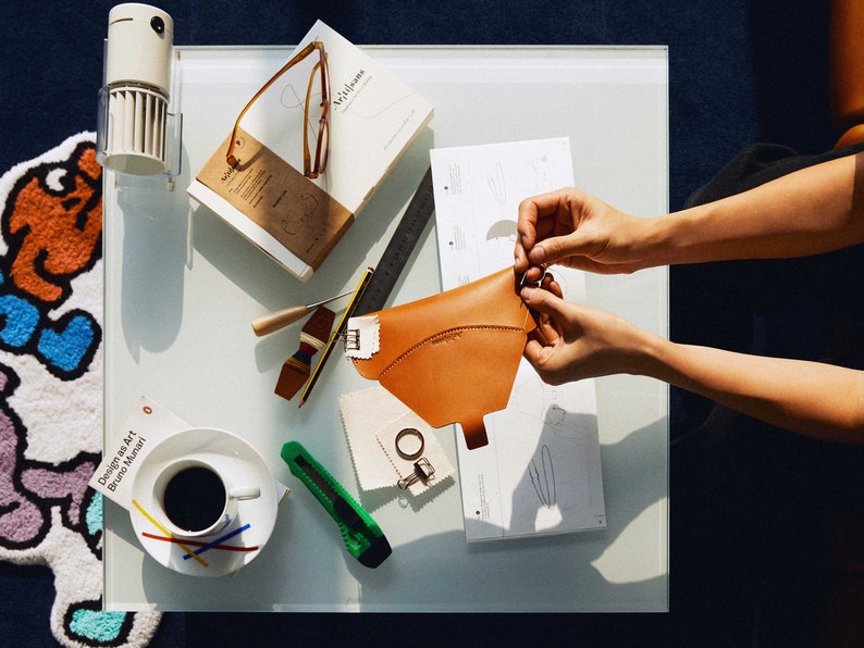 May include: A person working on a leather craft project. They are holding a brown leather piece and a pattern on a white surface. There are other tools and supplies on the table, including a book titled "Williams" and a cup of coffee.