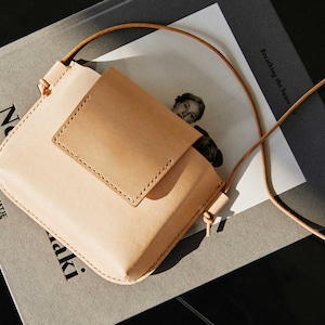 May include: A light brown leather crossbody bag with a flap closure and a small pocket on the front. The bag is sitting on a gray surface with a book underneath.