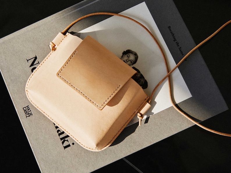 May include: A light brown leather crossbody bag with a flap closure and a small pocket on the front. The bag is sitting on a gray surface with a book underneath.