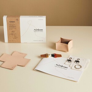 May include: A leathercraft kit with a box, a small container, a cross-shaped piece, a booklet, and other accessories. The box and container are light tan. The booklet has the text "Artisans" and "Experience the Art of Making".