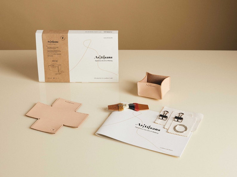 May include: A leathercraft kit with a box, a small container, a cross-shaped piece, a booklet, and other accessories. The box and container are light tan. The booklet has the text "Artisans" and "Experience the Art of Making".
