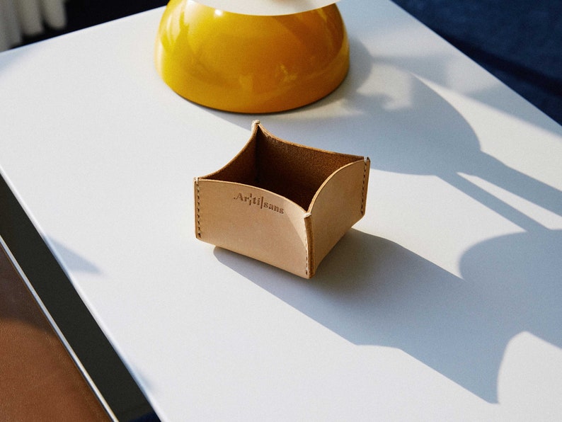 May include: A small, light brown leather tray with the word "Artisans" embossed on the front. The tray has a square shape with curved sides and is likely used for holding small items. A yellow lamp is in the background.