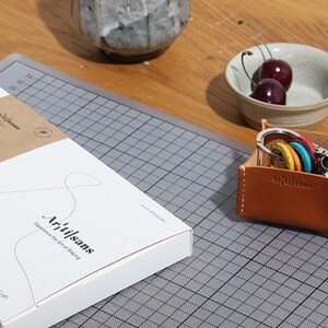 May include: A leather key holder with keys and a white box with the text "Artisans" and "Experience the Art of Making". The key holder is brown with white stitching and the keys have colorful keychains. Two cherries are in a small bowl.
