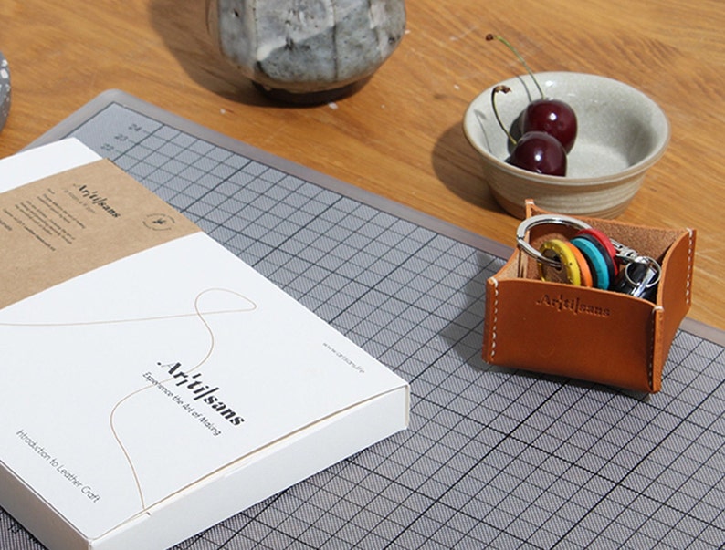May include: A leather key holder with keys and a white box with the text "Artisans" and "Experience the Art of Making". The key holder is brown with white stitching and the keys have colorful keychains. Two cherries are in a small bowl.