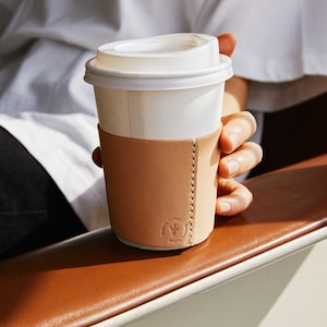 Op de afbeelding: Een witte koffiebeker om mee te nemen met een bruine leren hoes. De hoes heeft een logo met de tekst "Coffee Fuel Your Day".