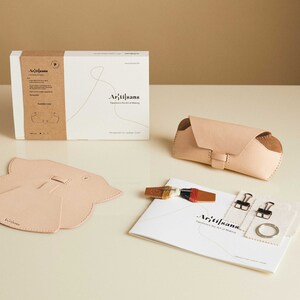 May include: A leather craft kit with all the materials needed to make a glasses case. The kit includes a pattern, leather pieces, thread, needles, and instructions. The kit is packaged in a brown box with the word "Artisans" printed on it.