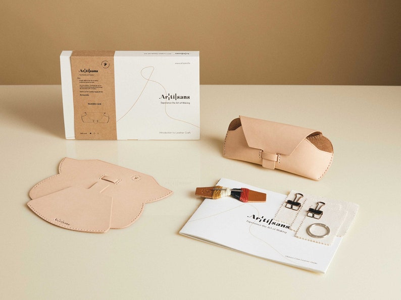 May include: A leather craft kit with all the materials needed to make a glasses case. The kit includes a pattern, leather pieces, thread, needles, and instructions. The kit is packaged in a brown box with the word "Artisans" printed on it.