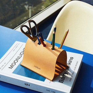 May include: A tan leather desk organiser with a curved shape sits on top of a white book with black text that reads "MOONLIGHT" and "Barry Jenkins". The organiser holds a pair of gold scissors, a blue pen, and three pencils.