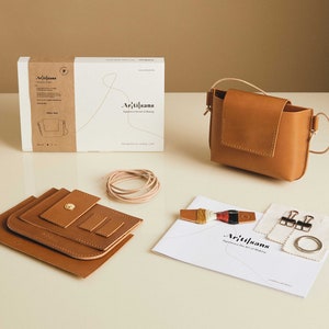 May include: A leather craft kit with instructions, tools, and materials to make a small brown leather bag. The kit includes a pre-cut leather bag, thread, a metal ring, and two metal clips.