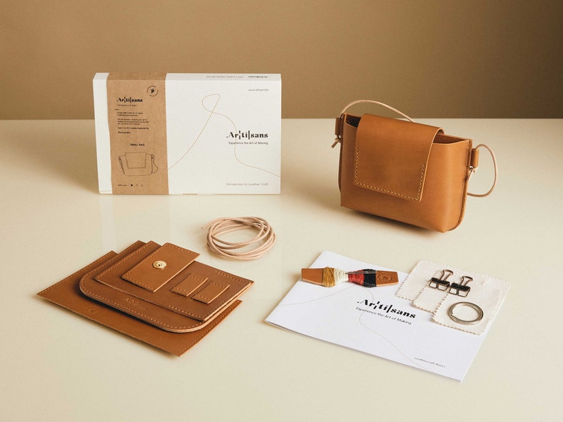 May include: A leather craft kit with instructions, tools, and materials to make a small brown leather bag. The kit includes a pre-cut leather bag, thread, a metal ring, and two metal clips.