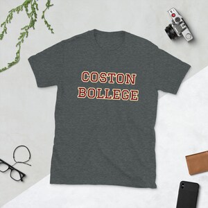 May include: Dark gray t-shirt with the text "COSTON BOLLEGE" in maroon letters with a cream-colored outline. The shirt is displayed on a flat surface with a camera, wallet, glasses, and a phone.