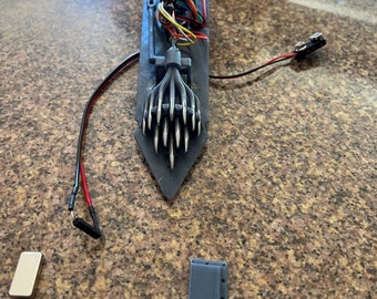 May include: A 3D printed gray robot arm with multiple metal claws. The arm is connected to a circuit board with wires. There are two gray rectangular blocks and a silver rectangular block on a brown surface.