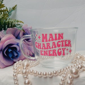 May include: A clear glass mug with a pink and white design that says "Main Character Energy". The mug is sitting on a white surface with a pearl necklace.
