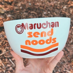 Könnte beinhalten: Weißer Keramiknapf mit dem Text "Maruchan send noods" in Orange und Braun. Die Schüssel hat ein lächelndes Gesichtslogo und orangefarbene und braune Wellenlinien. Die Schüssel wird in einer Hand gehalten.