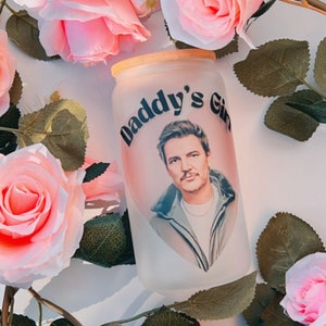 Puede incluir: Un vaso de vidrio esmerilado con fondo blanco y texto negro que dice "Daddy's Girl". El vaso presenta una imagen a color de un hombre con cabello castaño y barba que lleva una chaqueta azul. El vaso está rodeado de rosas rosas.