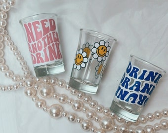Może przedstawiać: Trzy kieliszki do shotów z różnymi wzorami. Jeden z napisem "Need Another Drink" w różowych literach, drugi z motywem stokrotki z uśmiechniętą buźką w kolorze żółtym i białym, a trzeci z napisem "Drink Oranj" w niebieskich literach.