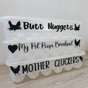 May include: Three clear plastic egg containers stacked on top of each other. Each container has a black graphic and text: "Butt Nuggets", "My Pet Poops Breakfast", and "MOTHER CLUCKERS". The containers are designed to hold eggs.