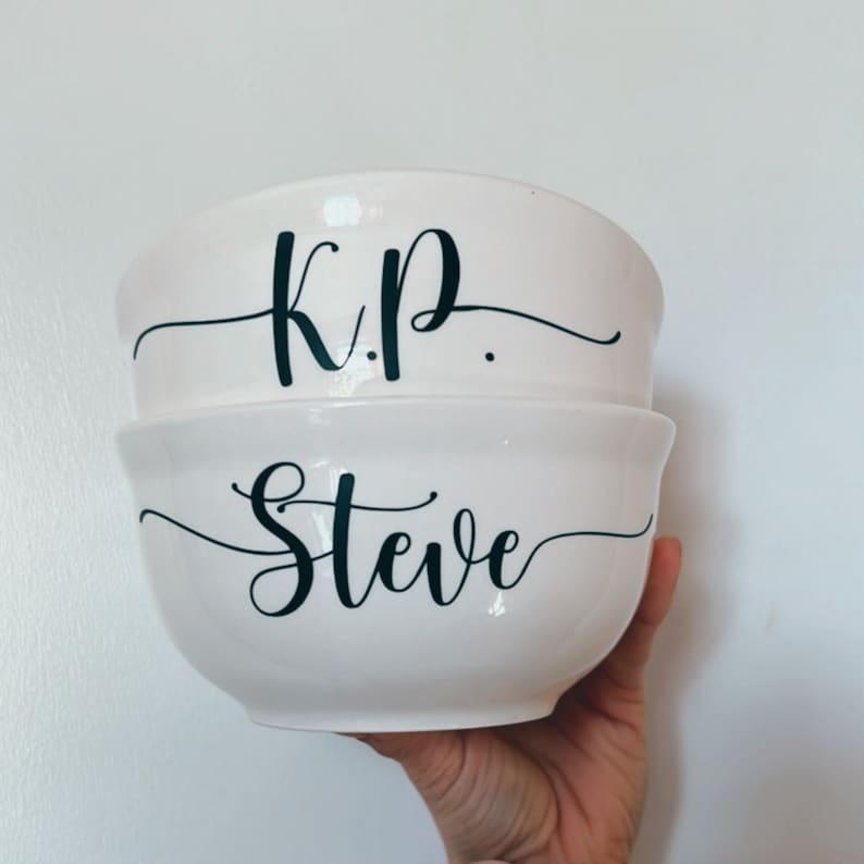 K&ouml;nnte beinhalten: Zwei wei&szlig;e Keramikschalen mit schwarzer kursivschrift. Die obere Schale sagt "K.P." und die untere Schale sagt "Steve".