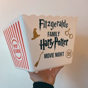 May include: White popcorn bucket with red and white stripes. The bucket has a black and gold design with the text "Fitzgeralds Family Harry Potter Movie Night 9 3/4".