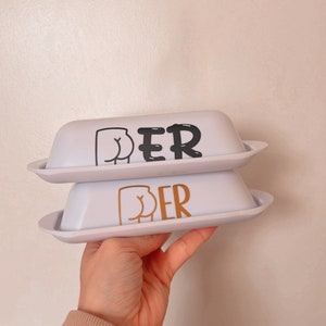 May include: Two white butter dishes with lids, stacked on top of each other. The top dish has black text and a black graphic of a butt. The bottom dish has gold text and a gold graphic of a butt.