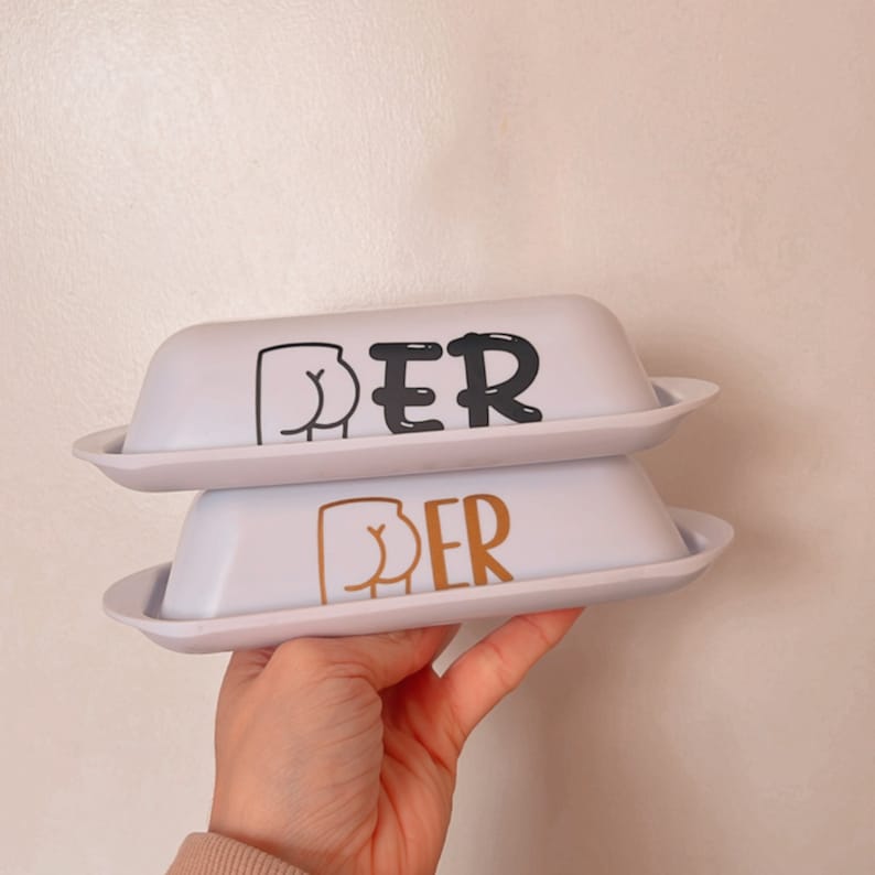 May include: Two white butter dishes with lids, stacked on top of each other. The top dish has black text and a black graphic of a butt. The bottom dish has gold text and a gold graphic of a butt.
