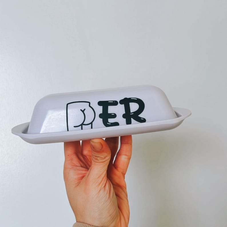 May include: A light gray butter dish with a lid, held by a hand. The dish has the word "BUTTER" printed on it in black, with the "BUT" replaced by a cartoon butt illustration. The dish is rectangular with rounded edges.