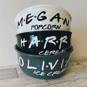 May include: Three stacked ceramic bowls with black and white lettering. The top bowl is white with the name "MEGAN" and the words "POPCORN" underneath. The middle bowl is black with the name "HARRY" and the word "CEREAL" underneath. The bottom bowl is gray with the name "OLIVIA" and the words "ICE CREAM" underneath.