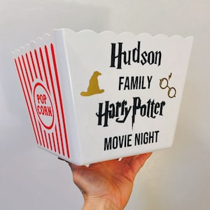 May include: White popcorn bucket with red and white stripes. The bucket has a black and gold design with the text "Hudson Family Harry Potter Movie Night".