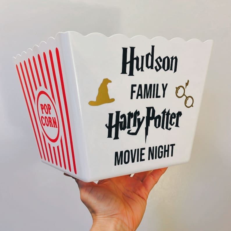 May include: White popcorn bucket with red and white stripes. The bucket has a black and gold design with the text "Hudson Family Harry Potter Movie Night".