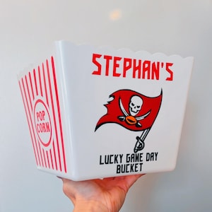 Puede incluir: Cubo de palomitas blanco con rayas rojas y negras. El cubo tiene un logotipo rojo y negro de los Tampa Bay Buccaneers y el texto "STEPHAN'S LUCKY GAME DAY BUCKET".