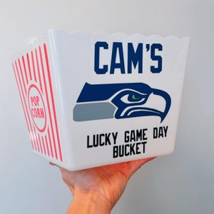 Puede incluir: Cubo de palomitas blanco con un logotipo de los Seattle Seahawks, las palabras "CAM'S LUCKY GAME DAY BUCKET" y un diseño a rayas rojas y blancas.