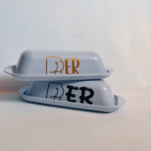 May include: Two light blue butter dishes stacked on top of each other. The top dish has a gold graphic of a butt and the word "PER" in gold. The bottom dish has a black graphic of a butt and the word "PER" in black.