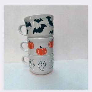 May include: Three white ceramic mugs stacked on top of each other. The top mug has a black bat design, the middle mug has an orange pumpkin design, and the bottom mug has a white ghost design.
