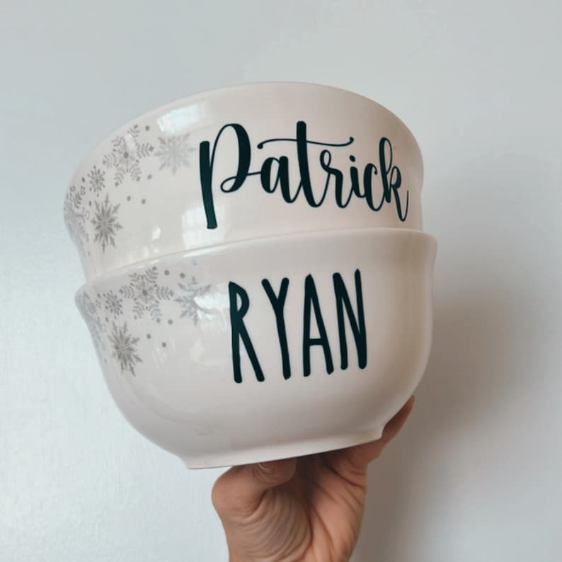 K&ouml;nnte beinhalten: Zwei wei&szlig;e Keramikschalen mit Schneeflockenmustern. Die obere Schale tr&auml;gt den Namen "Patrick" in schwarzer Schrift. Die untere Schale tr&auml;gt den Namen "Ryan" in schwarzer Schrift.