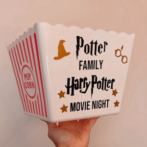 May include: White popcorn bucket with red stripes and a black and gold design. The bucket says "Potter Family Harry Potter Movie Night" with a wizard's hat, glasses, and stars.