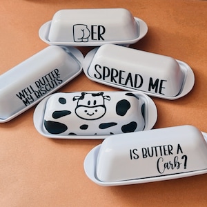 May include: A collection of white ceramic butter dishes with various black text and graphic designs. The designs include phrases like "SPREAD ME" and "IS BUTTER A CARB?" and a cow pattern.