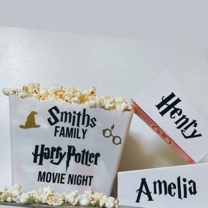 May include: White popcorn bucket with black text that reads "Smiths Family Harry Potter Movie Night". The bucket has a black silhouette of a witch's hat and a pair of glasses. Two white boxes with black text that reads "Henry" and "Amelia".