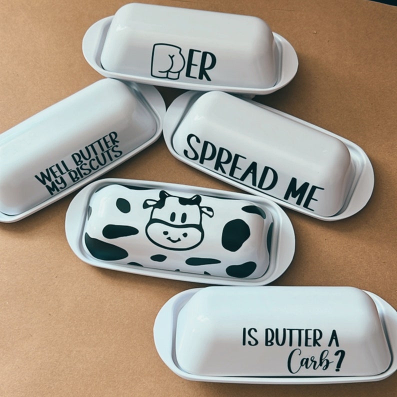 May include: Five white ceramic butter dishes with lids. One has a cow print, another says "Well Butter My Biscuits", and another says "Spread Me". The remaining two have the phrases "Is Butter a Carb?" and "Butter".