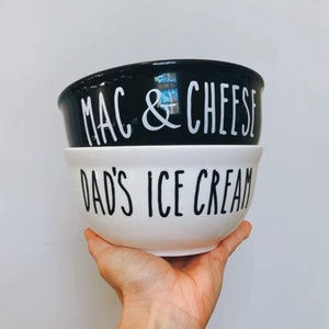 Può includere: Due ciotole in ceramica, una nera e una bianca, sovrapposte. La ciotola nera ha la scritta bianca "MAC & CHEESE". La ciotola bianca ha la scritta nera "DAD'S ICE CREAM".