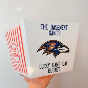以下が含まれることがあります： 赤いストライプと赤い円の中に「POP CORN」という文字が入った白いポップコーンバケツ。「THE BASEMENT GANG'S LUCKY GAME DAY BUCKET」という文字と、Bの文字が入ったカラスのロゴが入っています。