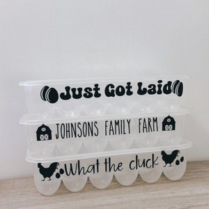 May include: Three clear plastic egg containers stacked on top of each other. Each container has a black graphic and text: "Just Got Laid", "Johnsons Family Farm", and "What the cluck". The containers are on a light-colored surface.