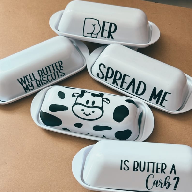 May include: White ceramic butter dishes with black text and designs. One dish has a cow pattern, another says "SPREAD ME", and others say "WELL BUTTER MY BISCUITS", "IS BUTTER A CARB?", and "BUTTER".