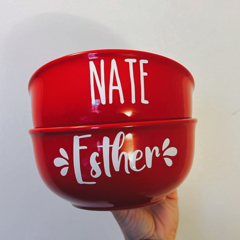 K&ouml;nnte beinhalten: Zwei rote Keramikschalen mit wei&szlig;er Beschriftung. Die obere Schale sagt "NATE" und die untere Schale sagt "Esther".
