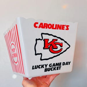 Può includere: Secchiello per popcorn bianco con strisce rosse e nere. Il secchiello ha un logo dei Kansas City Chiefs rosso e nero con il testo "CAROLINE'S" e "LUCKY GAME DAY BUCKET".