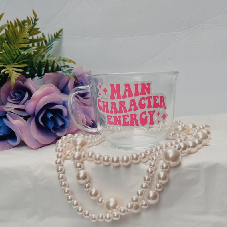 May include: A clear glass mug with a pink and white design that says "Main Character Energy". The mug is sitting on a white surface with a pearl necklace.